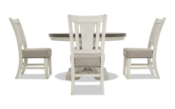 White wooden dining table set with beige upholstered cushioned chairs, featuring nailhead trim and a round pedestal table top.