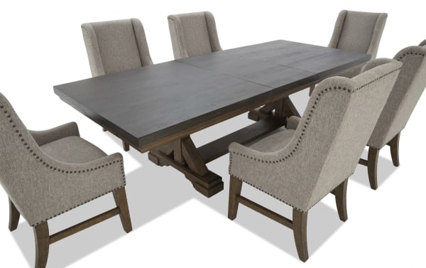 Rectangular dark wood dining table with trestle base, paired with six beige upholstered armchairs featuring nailhead trim and wooden legs.