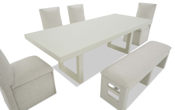 Modern white rectangular dining table set with cushioned beige fabric chairs and a matching bench, featuring geometric wooden legs.