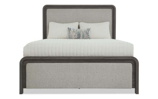 Queen-size bed with dark wood frame and light beige upholstered headboard and footboard, featuring white quilted bedding and decorative pillows.