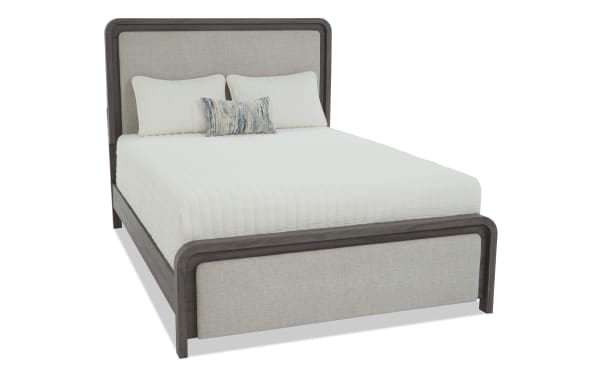 Queen-sized bed with gray wood frame and beige upholstered headboard and footboard, featuring a white quilted mattress and three pillows.