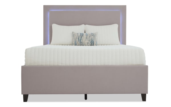 Modern upholstered bed with a light mauve fabric frame and headboard featuring built-in soft blue LED lighting, styled with white quilted bedding and decorative pillows.