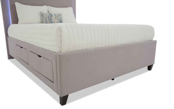 Upholstered platform bed with soft pink fabric, built-in side storage drawers, tapered dark wooden legs, and white quilted bedding with decorative pillows.