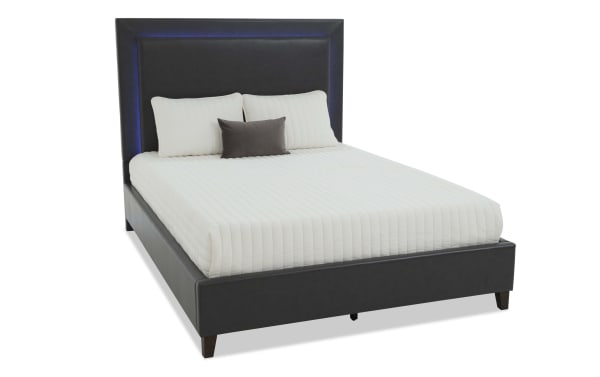 Modern black faux leather platform bed with tall padded headboard, white quilted mattress cover, and three pillows, suitable for bedroom decor.