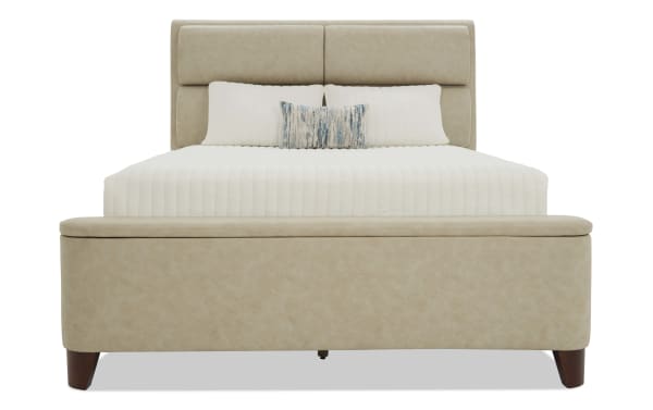 Beige upholstered bed with padded headboard and footboard, white quilted bedding, decorative blue and beige accent pillow, wooden legs, modern style.