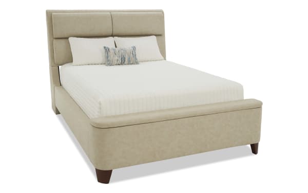 Beige upholstered bed with padded headboard and footboard, wooden legs, white quilted bedding, and decorative pillows, modern bedroom furniture.