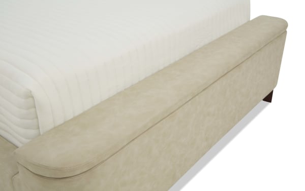 Beige upholstered bed bench with rounded edges and wooden legs, placed at the foot of a white textured mattress.