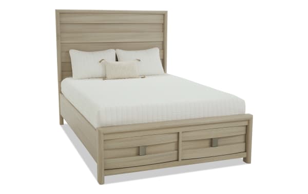 Queen-size wooden bed frame in light oak finish with a tall paneled headboard, footboard storage drawers, and white quilted bedding with pillows.