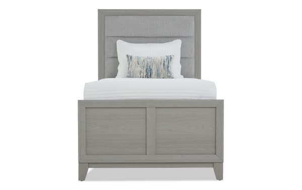 Twin bed with light gray wooden frame and upholstered headboard, featuring white bedding and a decorative blue and beige accent pillow.