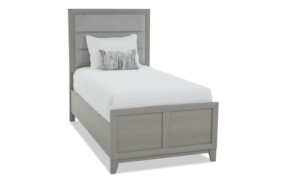 Twin bed with a light gray wooden frame and upholstered gray fabric headboard, featuring white bedding and decorative pillows, modern style.