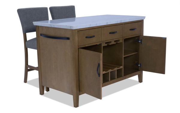 Wooden kitchen island with white marble countertop, three drawers, two open cabinet doors revealing wine rack and shelves, paired with two gray upholstered bar stools.