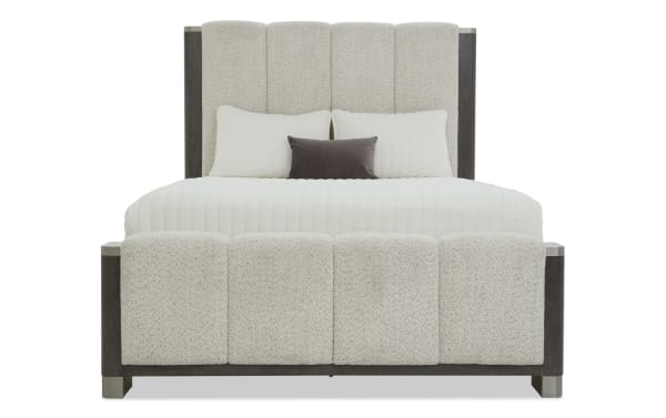 Modern upholstered bed with a tall, tufted beige fabric headboard and matching footboard, featuring dark wood accents and white bedding with decorative pillows.