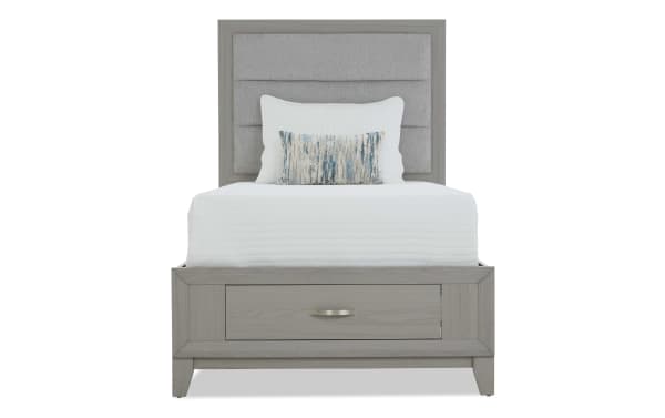 Twin bed with gray wooden frame, upholstered padded headboard, white bedding, and decorative pillows, featuring a storage drawer at the base.