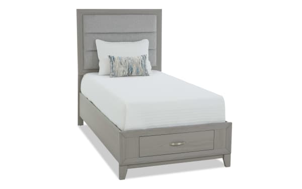 Twin bed with gray wood frame and cushioned gray fabric headboard, featuring a storage drawer at the foot, styled with white bedding and decorative pillows.