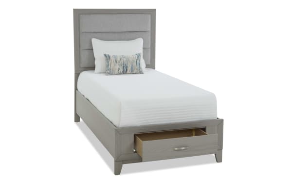 Twin bed with light gray wooden frame and upholstered headboard, featuring a built-in storage drawer and white bedding with decorative pillows.