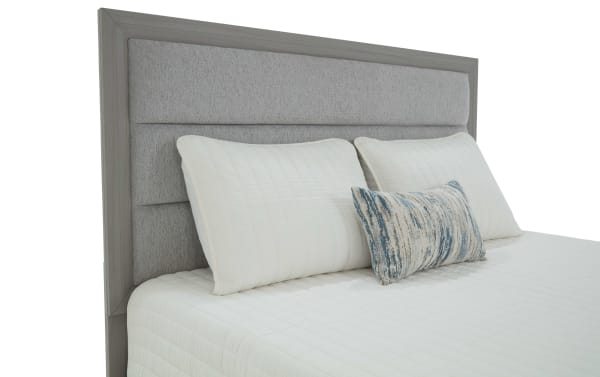 King-size bed with a grey upholstered headboard featuring horizontal padded panels, white quilted bedding, two large white pillows, and a decorative blue and beige lumbar pillow.