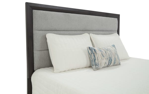 King-size bed with a dark wood frame and a light gray upholstered headboard, featuring white quilted pillows and a decorative blue-and-gray accent pillow.