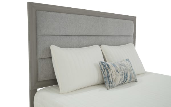 Gray wooden bed frame with a padded, textured gray fabric headboard, featuring white quilted pillows and a small decorative blue and beige accent pillow.