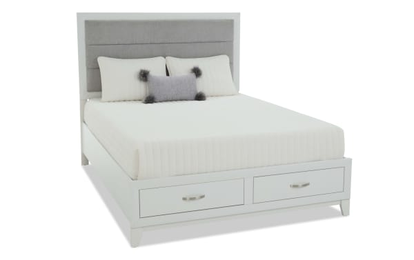 Modern white wooden bed frame with gray upholstered headboard, white quilted bedding, two white pillows, and a small decorative gray pillow with black pom-poms.