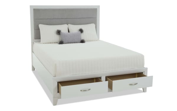 White wooden bed frame with gray upholstered headboard, white quilted bedding, decorative pillows, and two open storage drawers at the foot.