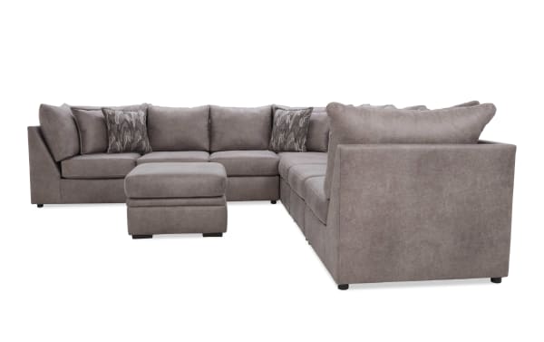 Modern L-shaped sectional sofa in taupe microfiber fabric with plush cushions, two patterned accent pillows, and matching ottoman for living room seating.