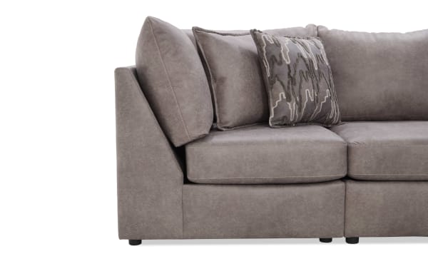 Section of a modern taupe microfiber sectional sofa with angled armrest, plush seat cushions, and three decorative throw pillows including one with a gray abstract pattern.