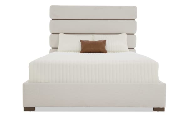 Modern upholstered bed with a beige fabric frame and headboard featuring horizontal panel design, white quilted bedding, and a brown accent pillow.