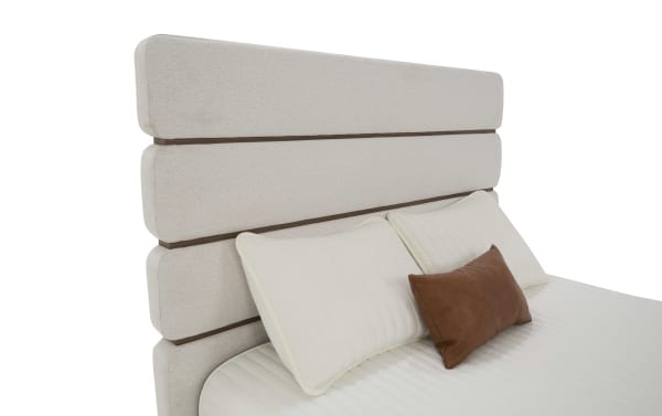 Modern beige upholstered bed headboard with horizontal wooden accents, paired with white quilted pillows and a brown leather lumbar pillow.