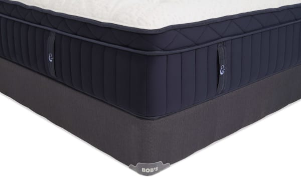 Corner view of a navy blue and white mattress with quilted top and vertical stitching on sides, resting on a dark gray fabric foundation labeled "BOB'S."