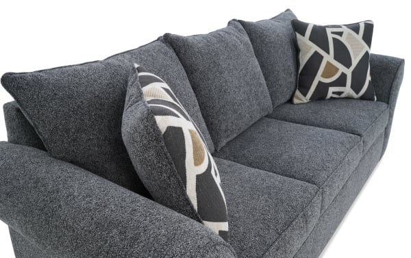 Dark gray fabric sofa with textured upholstery, three large seat cushions, two patterned accent pillows in black, beige, and brown geometric design, and wide armrests.