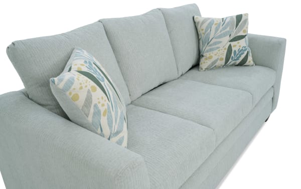 Light blue fabric sofa with three seat cushions and three back cushions, featuring two decorative leaf-patterned throw pillows in blue, green, and yellow tones.