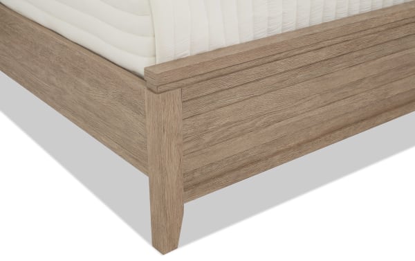 Corner view of a wooden bed frame in a light oak finish with a white mattress, showcasing the textured wood grain and sturdy leg design.