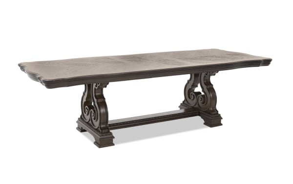 Dark brown wooden dining table with ornate carved pedestal legs, rectangular top with beveled edges, and a connecting stretcher bar.