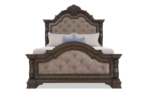 King-size bed with dark wood frame and ornate carved details, featuring tufted beige upholstered headboard and footboard, styled with neutral bedding and a blue accent pillow.