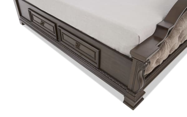 Dark wood bed frame corner with carved decorative details, two storage drawers, and upholstered side panel in beige fabric.