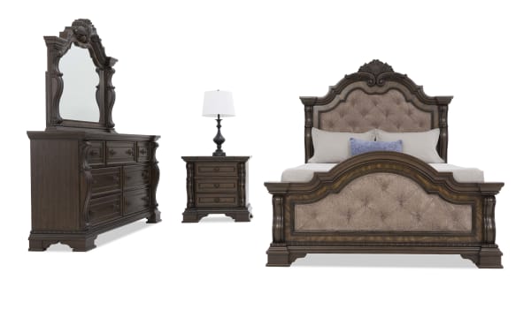 Traditional dark wood bedroom set with ornate carved details, featuring a tufted upholstered headboard and footboard, matching dresser with mirror, and nightstand with lamp.