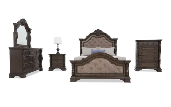 Traditional dark wood bedroom set with ornate carved details, featuring a tufted upholstered bed, matching dresser with mirror, nightstand, and chest.