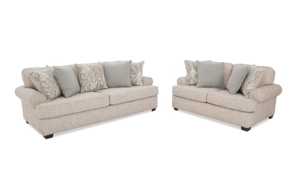 Two beige fabric sofas with rolled arms and dark wooden legs, each adorned with patterned and solid decorative throw pillows, ideal for living room seating.