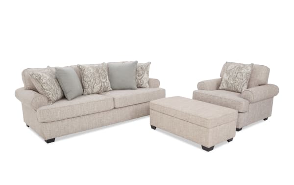 Beige fabric living room set featuring a three-seater sofa, matching armchair, and rectangular ottoman, all with rolled arms and decorative throw pillows.