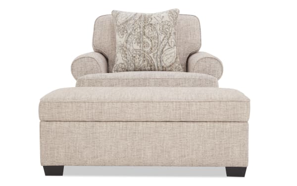 Beige upholstered armchair with matching ottoman, featuring a textured fabric, rounded arms, cushioned back, and decorative paisley-patterned throw pillow.