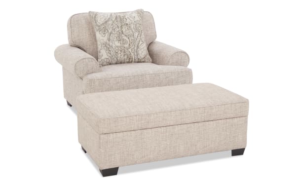 Beige fabric armchair with rolled arms and matching ottoman, featuring a decorative paisley-patterned throw pillow for added comfort and style.