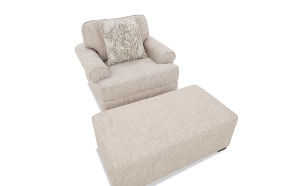 Beige upholstered armchair with rolled arms and a matching rectangular ottoman, featuring a decorative patterned throw pillow.