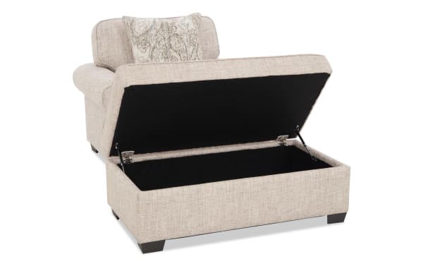 Beige fabric storage ottoman with hinged lid open, showing spacious black interior, dark wooden legs, and decorative pillow on top.