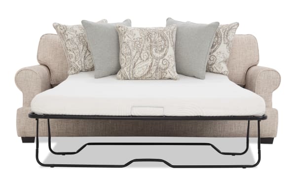 Beige fabric sofa bed with rolled arms, featuring a white mattress and five decorative pillows in beige paisley and solid gray patterns, supported by a black metal frame.