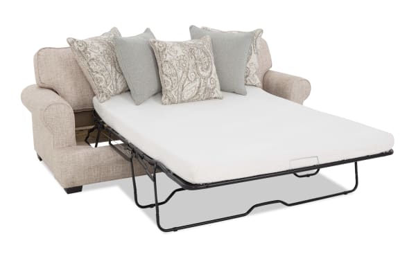 Beige fabric loveseat sleeper sofa with a white mattress and four decorative pillows, featuring a fold-out metal frame bed for space-saving functionality.