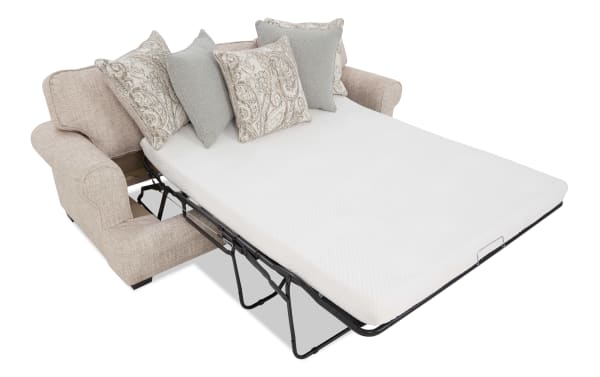 Beige fabric sofa bed with a fold-out metal frame and white mattress, featuring decorative patterned and solid color throw pillows.
