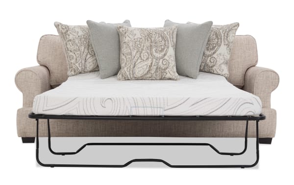 Beige fabric sofa bed with rolled arms, featuring a black metal frame fold-out mattress with a white quilted cover and five decorative throw pillows in beige and gray patterns.