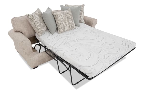 Beige fabric sofa bed with a white patterned mattress and decorative pillows, featuring a fold-out metal frame for sleeping.