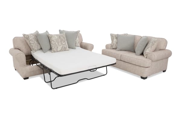Beige fabric sectional sofa with a pull-out metal frame sleeper bed, including a white mattress and decorative patterned and solid gray cushions.
