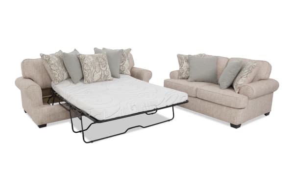 Beige fabric sleeper sofa with rolled arms and dark wooden legs, featuring a pull-out metal frame bed with a white patterned mattress and decorative paisley and solid gray cushions.
